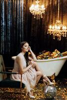 Beautiful and stylish young girl in a dress sitting near a bathtub with gifts on a black background photo