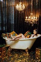 A young and beautiful girl is lying in the bath with New Year's gifts in golden wrappers on a dark background photo