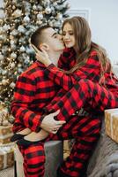 A young beautiful married couple in the same red pajamas, near the Christmas tree, are smiling, hugging and kissing. New Year holidays and gifts under the Christmas tree photo