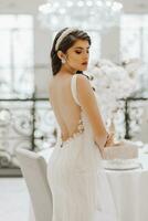 Beautiful bride at the table with a vase of flowers. The woman is wearing a long wedding dress with lace and an open back, a bow is tied on the back. photo