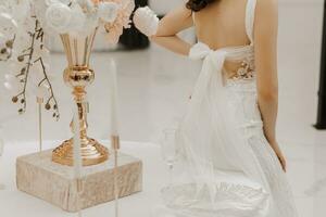 Beautiful bride at the table with a vase of flowers. The woman is wearing a long wedding dress with lace and an open back, a bow is tied on the back. photo