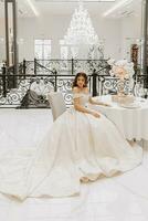 studio photo of a girl in a wedding dress with a luxurious hairstyle and makeup, the bride is sitting at the table