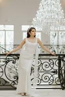 studio photo of a girl in a wedding dress near a black metal railing. A large hanging chandelier. Luxury interior in white style