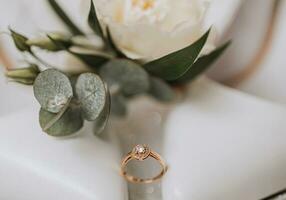 A gold wedding ring with a diamond near a rose with greenery, on a white background. Wedding details in the photo. photo