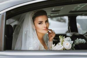 A brunette bride in a lace dress and voluminous veil in the car window. A beautiful and smiling bride. Happy weekend. photo