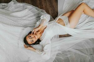 A brunette bride is dressed in a satin robe, posing while lying on the floor. Bare legs. Beautiful hair and make-up, open bust. Wedding portrait. Gorgeous silver jewelry photo