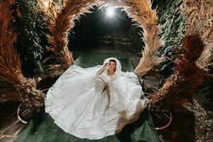 Portrait of the bride. The bride is sitting against the background of an arch made of ears of corn. Long train of the dress. Voluminous veil. Crystal jewelry. Rustic style photo