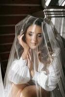 el novia es preparando para el boda. un soñador niña en un vendaje vestido con largo oscuro pelo cerca el ventana debajo un velo foto