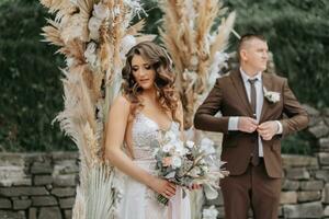 Portrait of the bride and groom. The bride stands against the background of the groom and an arch made of flowers and dried flowers. Stylish wedding dress. Wedding decorations. Rural style photo