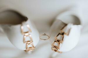 An elegant gold wedding ring with a diamond, standing between women's white high-heeled shoes. Photo from above on a white background