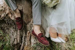 recortado foto de el novia y novio sentado en un árbol tocón en el bosque en elegante atuendo. ramo de flores de flores desde peonias, rojo Zapatos de el novio