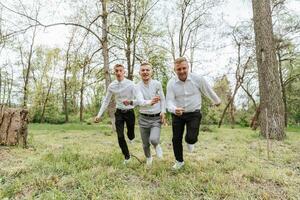 The groom and his friends pose against the background of the forest. not a large company of people having fun. Smiling boys run through the forest and rejoice together with the bridegroom photo