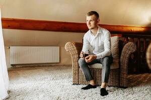 A young businessman in a hotel room sits on a chair and drinks his whiskey from a glass. Rest in wonderful apartments. Groom's morning at the hotel photo