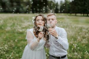 the bride and groom walk in the field on their wedding day and blow dandelions into the camera photo