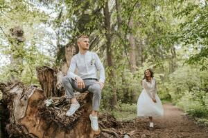 Wedding walk in the forest. The bridegroom is seated on a felled old tree in the foreground. Country wedding concept. photo