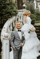 retrato de el novia y novio. el novia en un Boda vestir con un tren en un elegante escalera. novio en un clásico gris traje, blanco camisa y arco atar. lleno longitud foto. Boda en un parte superior ubicación foto