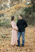 retrato de un contento joven familia, marido y embarazada esposa, en pie en contra el antecedentes de hermosa ligero en el otoño bosque, mirando a cada otro foto