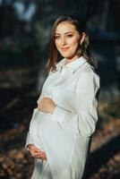 A pregnant woman in a white dress on a dark background poses in the park. Happy and carefree pregnancy. Autumn park. Happy woman enjoying her pregnancy. photo