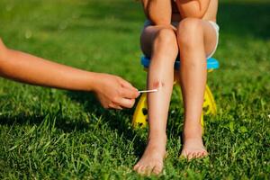 A woman's hand treats a child's wound under the knee with a disinfectant. A child sits on a chair and green grass in the summer. Treatment of the wound. First aid. Cropped photo