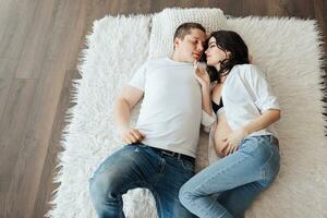 moderno hermosa embarazada mujer en pantalones y camisa con su marido en el cama. foto desde arriba. concepto de el embarazo, maternidad, preparación y espera. el belleza de un mujer durante el embarazo