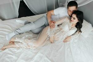 Young brunette pregnant woman in her room in a white lace dress on the bed with her husband. Motherhood. Concept of pregnancy, motherhood, preparation and waiting. photo