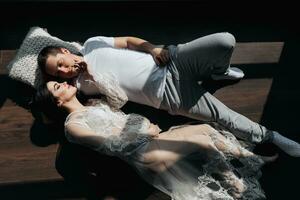 photo of a young pregnant woman in her room in a white lace dress on the floor with her husband in beautiful sunlight. Motherhood. Concept of pregnancy, motherhood, preparation and waiting.