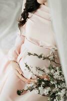 close up of a beautiful pregnant woman with long dark hair in a pink dress touching her belly while sitting on the bed photo