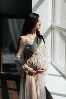 a beautiful pregnant woman in a lace transparent dress hugs her tummy with her hands near the window. Concept of pregnancy, motherhood, preparation and waiting. The beauty of a woman during pregnancy photo