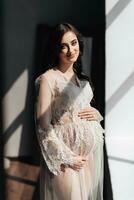 a beautiful pregnant woman in a lace transparent dress hugs her tummy with her hands near the window. Concept of pregnancy, motherhood, preparation and waiting. The beauty of a woman during pregnancy photo