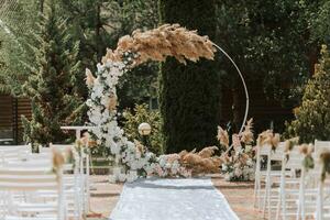 The wedding arch in the park is made of fresh flowers and dry reeds. Away wedding ceremony. White chairs for the ceremony photo