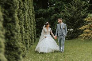 European wedding couple walking in the park. The bride in a beautiful dress with sleeves and a crown on her head. Groom with a black beard of Caucasian appearance in a classic suit. photo