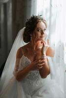 Portrait of a beautiful girl with brown hair in a white openwork dress with a wreath of flowers on her head, standing behind the window photo