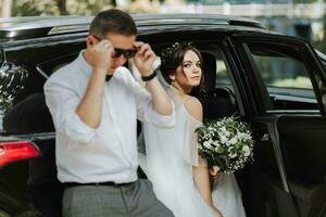 Boda retrato, foto de un elegante novio en un blanco camisa y arco Corbata y un morena novia con un ramo de flores de flores cerca un negro coche.