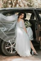 A beautiful bride in a white dress with a bouquet poses near a car in nature. Wedding portrait of a cute girl. Photography and concept. photo