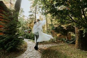 Fashionable groom and cute bride in white dress with tiara of fresh flowers circling and laughing in park, garden, forest outdoors. Wedding photography, portrait of smiling newlyweds. photo