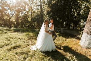 un joven Pareja de recién casados son caminando en un verano parque. el novia en un hermosa blanco vestir con un guirnalda de Fresco flores en su cabeza, el novio en un blanco camisa foto