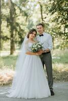 Fashionable groom and cute bride in white dress with tiara of fresh flowers, hugging, laughing in park, garden, forest outdoors. Wedding photography, portrait of smiling newlyweds. photo