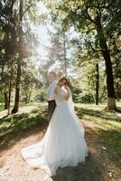 Fashionable groom and cute bride in white dress with tiara of fresh flowers, hugging, laughing in park, garden, forest outdoors. Wedding photography, portrait of smiling newlyweds. Wide angle photo