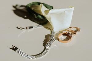 a boutonniere of calla flowers, a gold wedding ring on the table, a silver bracelet photo