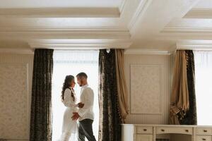 Beautiful, sexy bride in a white robe, groom in a white shirt hugs the bride in a hotel room. Wedding portrait of newlyweds in love. Free space. photo