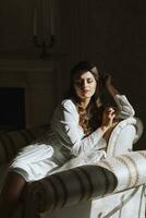 Portrait of a young attractive brunette with long hair in a white robe. She is sitting on a chair in the hotel. A dream bride. photo