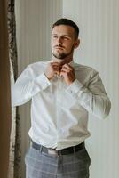 A stylish groom with a beard is preparing for the wedding ceremony. Groom's morning. A businessman is buttoning up a white shirt. The groom is getting ready in the morning before the wedding ceremony photo