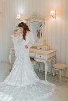 A beautiful young bride in a white petticoat is standing and holding a wedding dress in her hands. Final preparations for the wedding. The bride is waiting for the groom. Morning, bride. photo