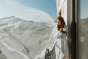 A beautiful bride in a white robe drinks coffee from a white cup on a hotel balcony overlooking a big city. On your wedding day before marriage. photo