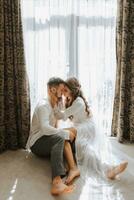 Beautiful, sexy bride in a white robe, groom in a white shirt hugs the bride against the background of the window in the hotel room. Wedding portrait of newlyweds in love. Vertical photo. photo