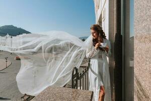 A beautiful bride in a white robe drinks coffee from a white cup on a hotel balcony overlooking a big city. On your wedding day before marriage. photo