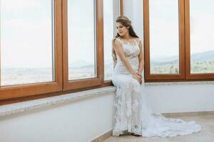 the bride in an evening dress sits on the windowsill against the background of large windows. Beautiful young bride. Free space photo