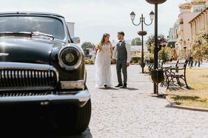 frente ver de un casado novia y novio vestido en festivo ropa caminando mediante el ciudad a su retro auto, participación un Boda ramo, sonriente y mirando a cada otro en su Boda día foto