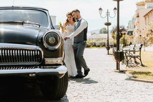 Front view of married bride and groom wearing festive clothes walking through the city to their retro car, holding wedding bouquet, smiling on wedding day photo