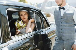 Wedding portrait photo of stylish groom in white shirt, waistcoat and bow tie and brunette bride with bouquet of flowers near black retro car.